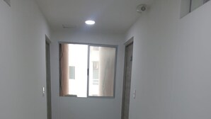 Un pasillo estrecho con una ventana al final, paredes blancas y suelo de baldosas.