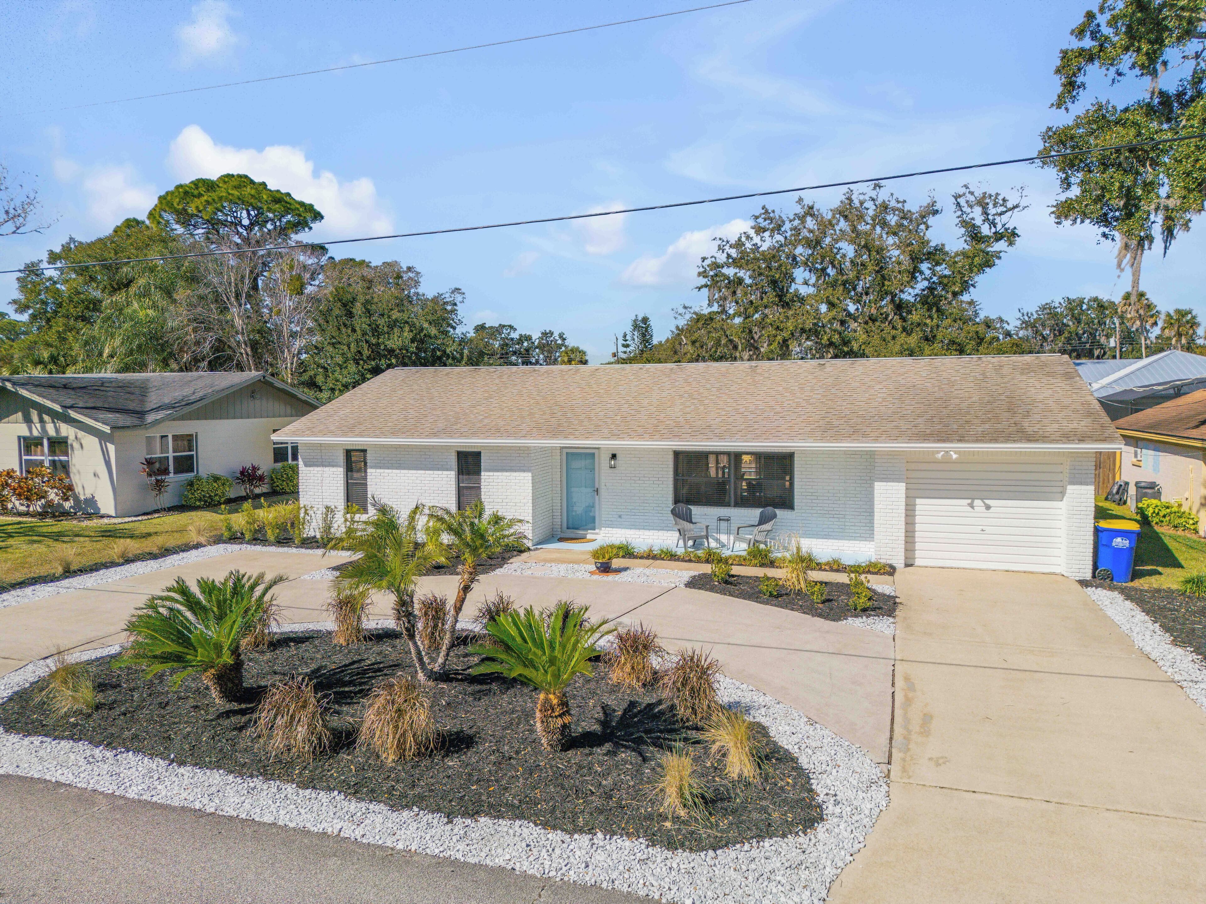 A single-story house with a front porch, a garage, and a landscaped front yard with palm trees and shrubs.