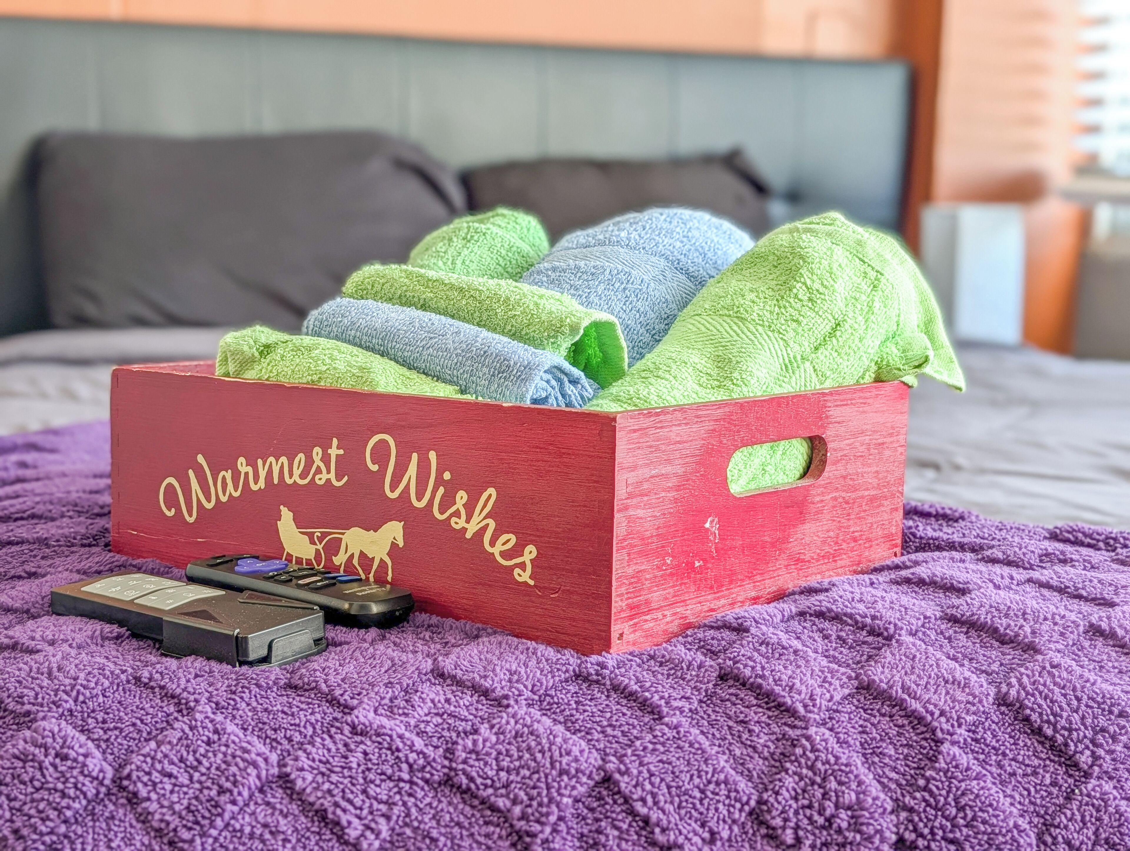 Caixa vermelha com a etiqueta 'Warmest Wishes', com toalhas coloridas e um comando remoto sobre um cobertor roxo.