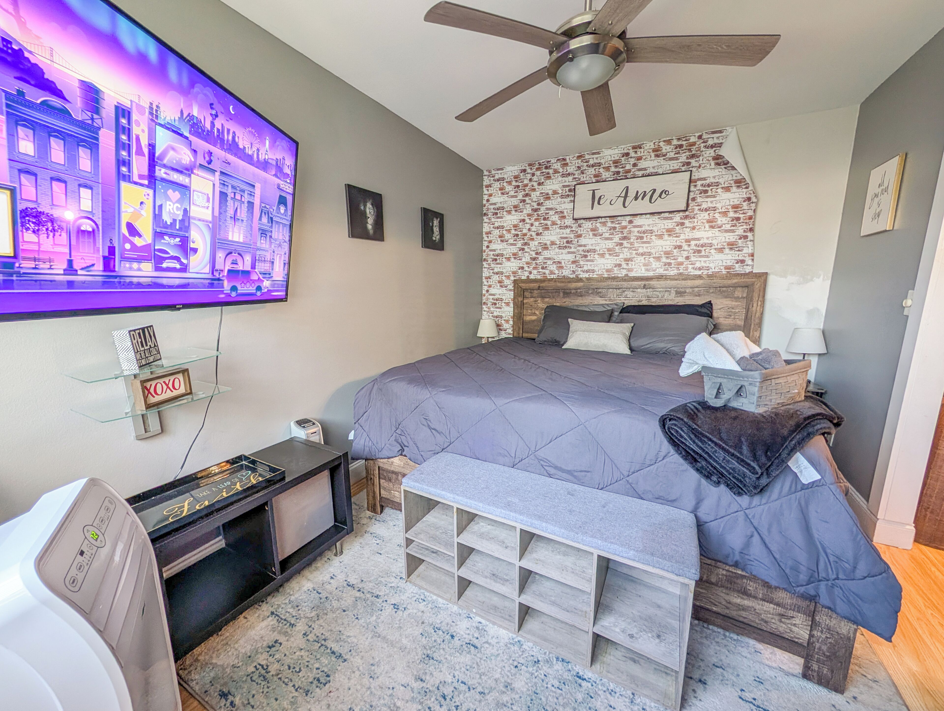 A bedroom with a bed, a ceiling fan, a TV, a brick-patterned wall, and a sign that says 'Te Amo'.