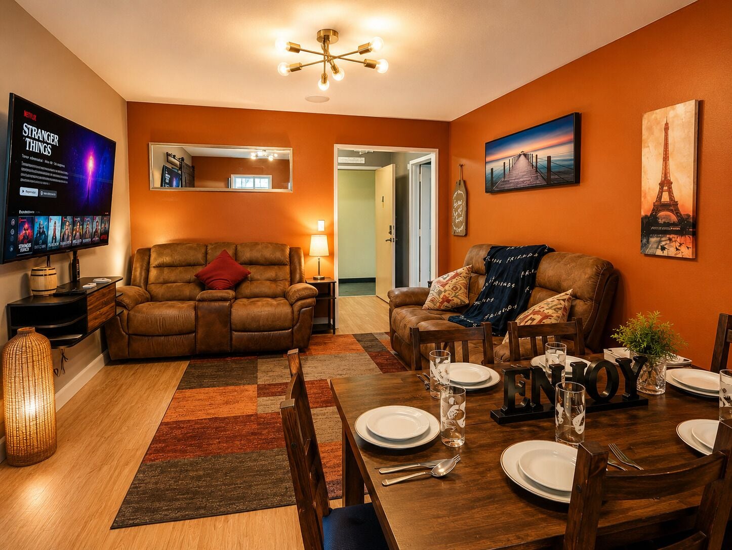 Una sala de estar con televisor, sofás, una mesa de comedor preparada para cuatro personas y un pasillo que lleva a otra habitación.
