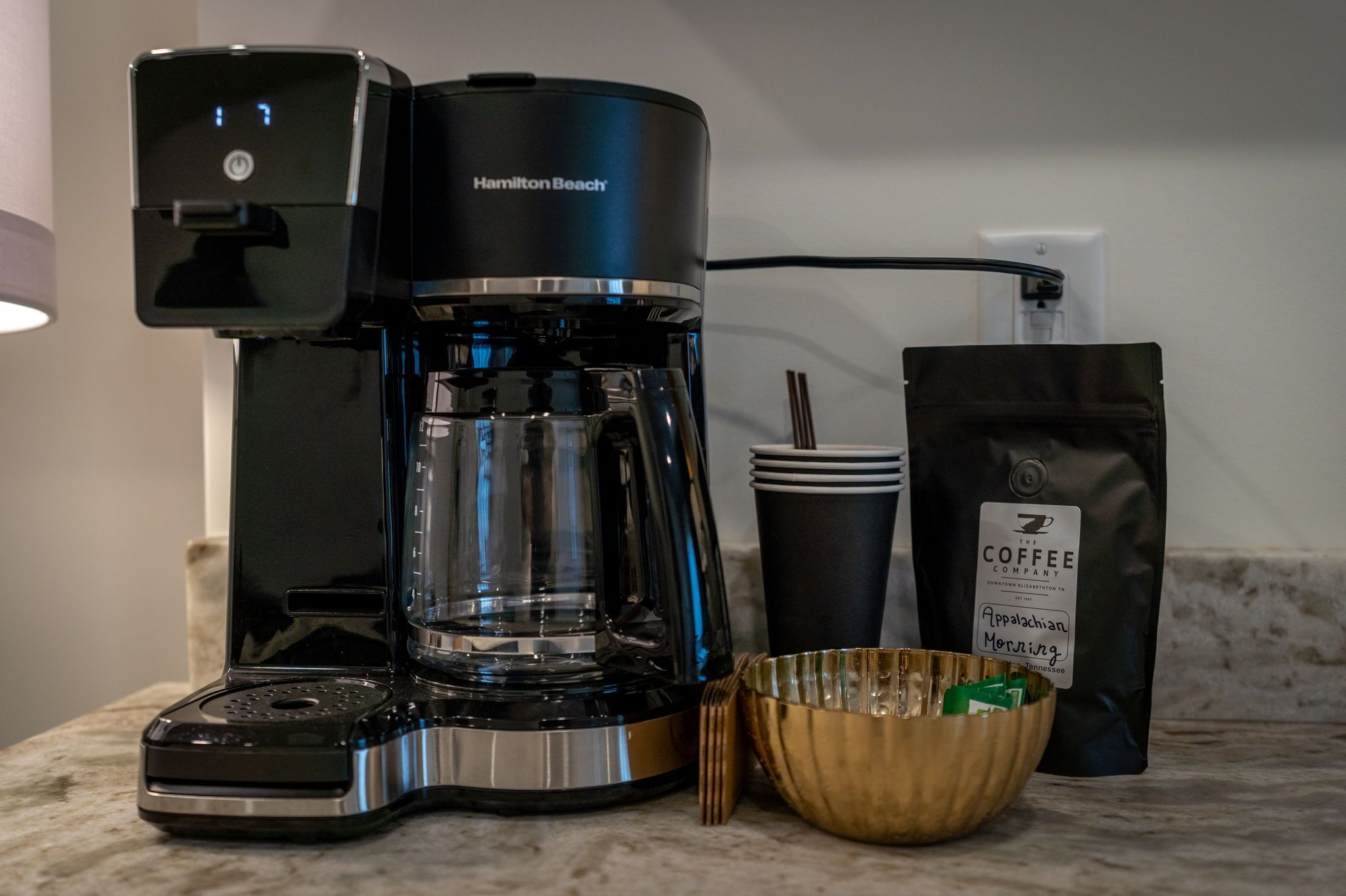 A Hamilton Beach coffee maker, a coffee cup, a bag of coffee, and a bowl on a kitchen counter.