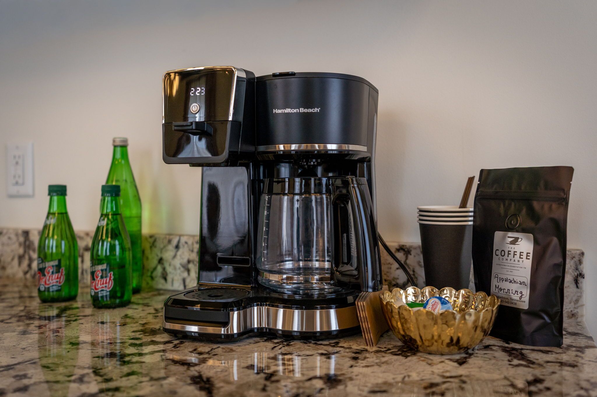 Una cafetera, botellas de refresco verde, una bolsa de café y un bol sobre una encimera de cocina.
