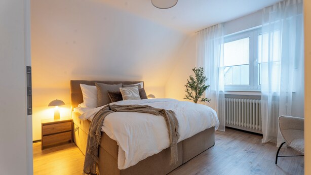 Ein Schlafzimmer mit Bett, Nachttisch, Lampe, Fenster und Stuhl.