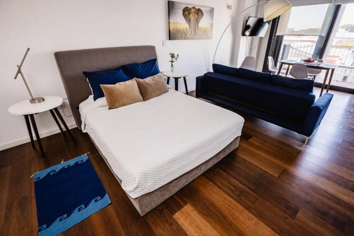 A modern bedroom with a large bed, a dark blue sofa, a small round table, and a wall-mounted TV.