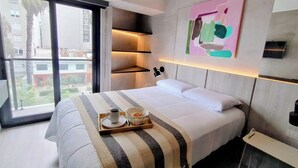 Une chambre d’hôtel moderne avec un grand lit, une table de chevet, un plateau avec une tasse et un sac, un tableau au mur et une vue sur les bâtiments par la fenêtre.