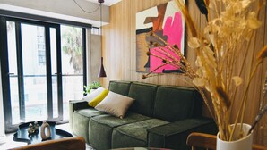 Un salon moderne avec un canapé vert, une table basse en verre et une grande fenêtre donnant sur les bâtiments environnants.