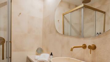 Un baño moderno con un lavamanos ovalado grande, cabina de ducha con mampara de vidrio y un espejo con marco dorado.