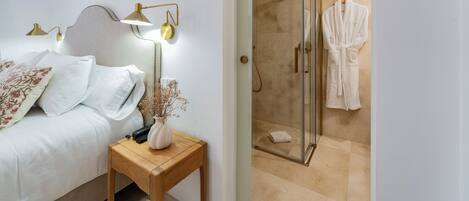 A bedroom with a bed, bedside table, lamp, and a glass-enclosed shower area with a robe hanging on the wall.