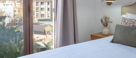 A hotel room with a large bed, a nightstand, a lamp, a vase with dried flowers, and a view of buildings and hills.