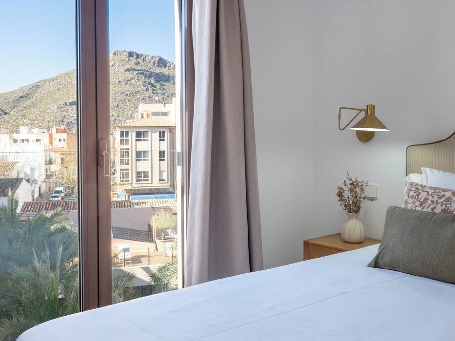 Habitación de hotel con una cama grande, una mesita de noche, una lámpara, un jarrón con flores secas y vistas a edificios y colinas.