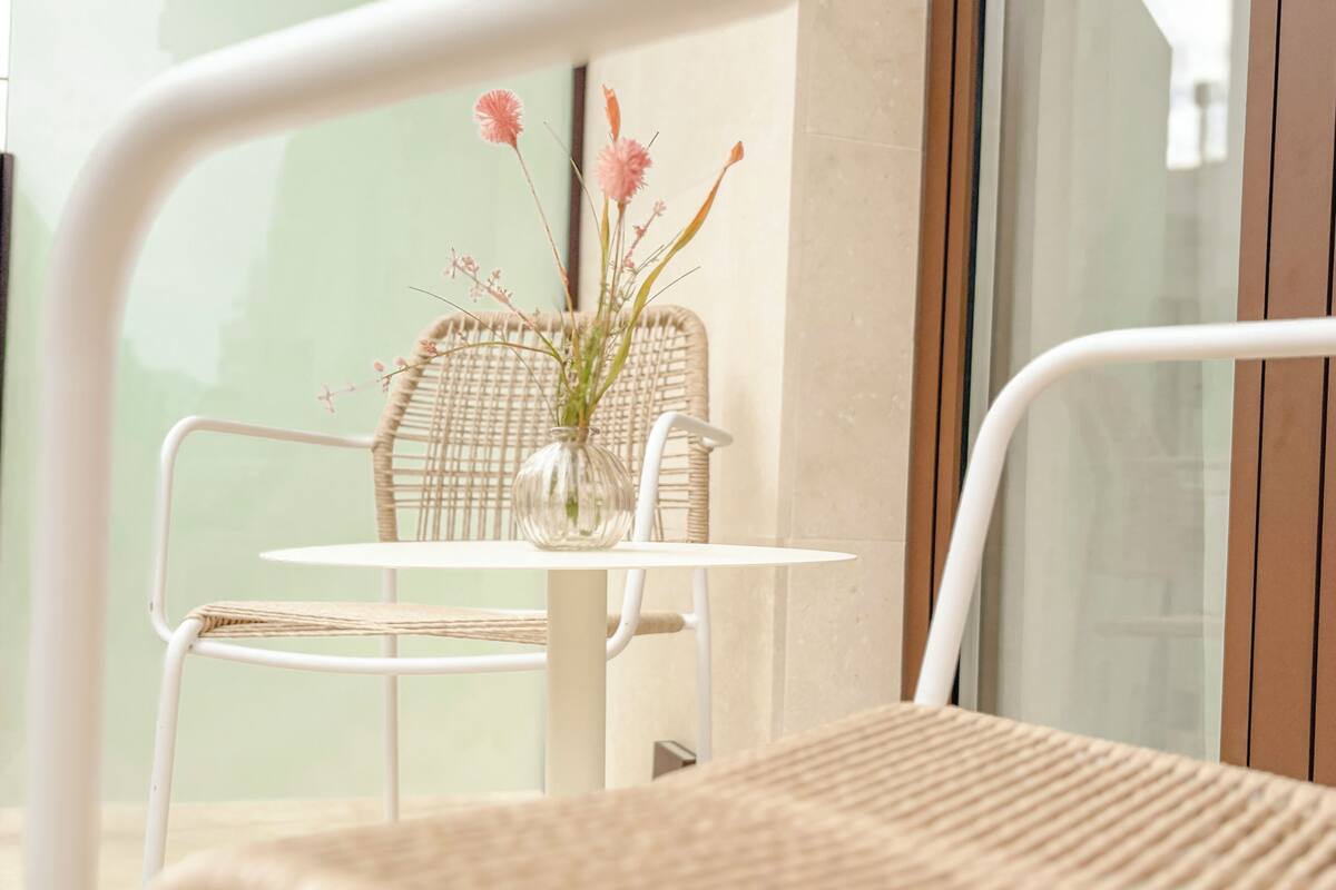 A white metal chair with a woven seat and backrest, a small round table, and a clear glass vase with pink flowers.