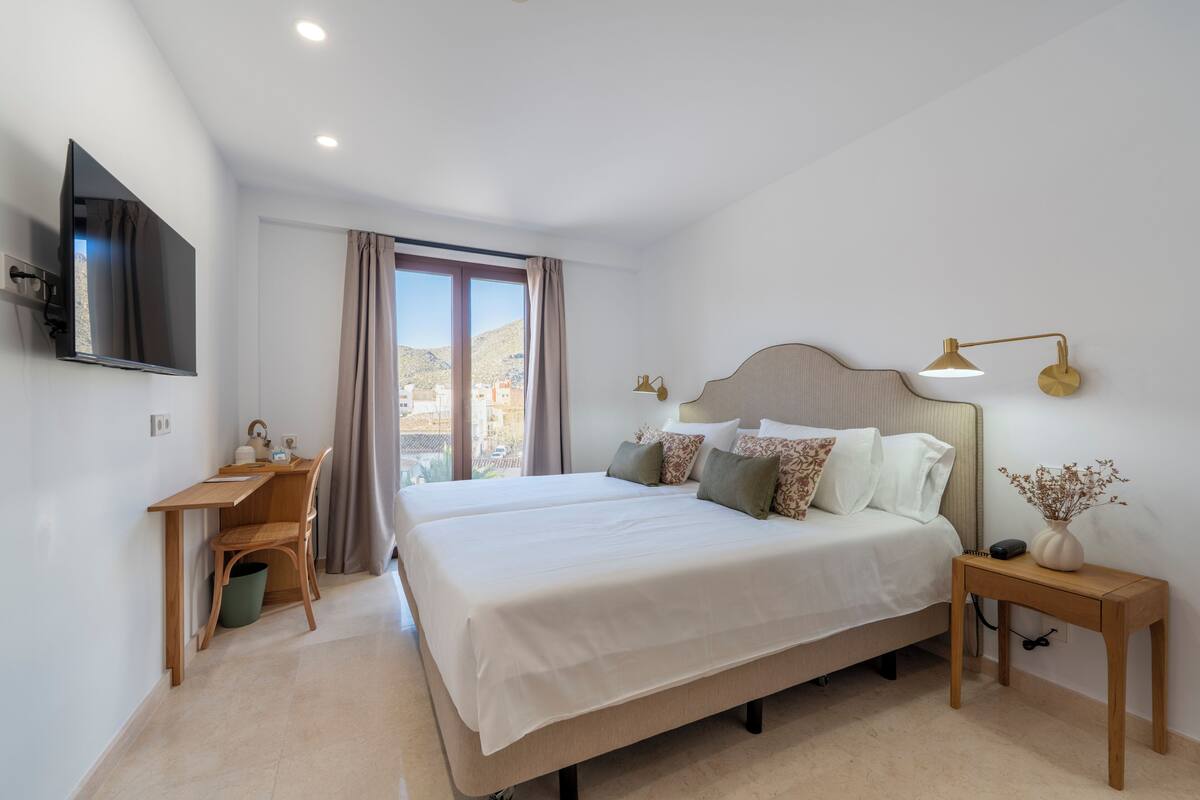 A bedroom with a bed, a desk, a chair, a TV, a lamp, and a window.