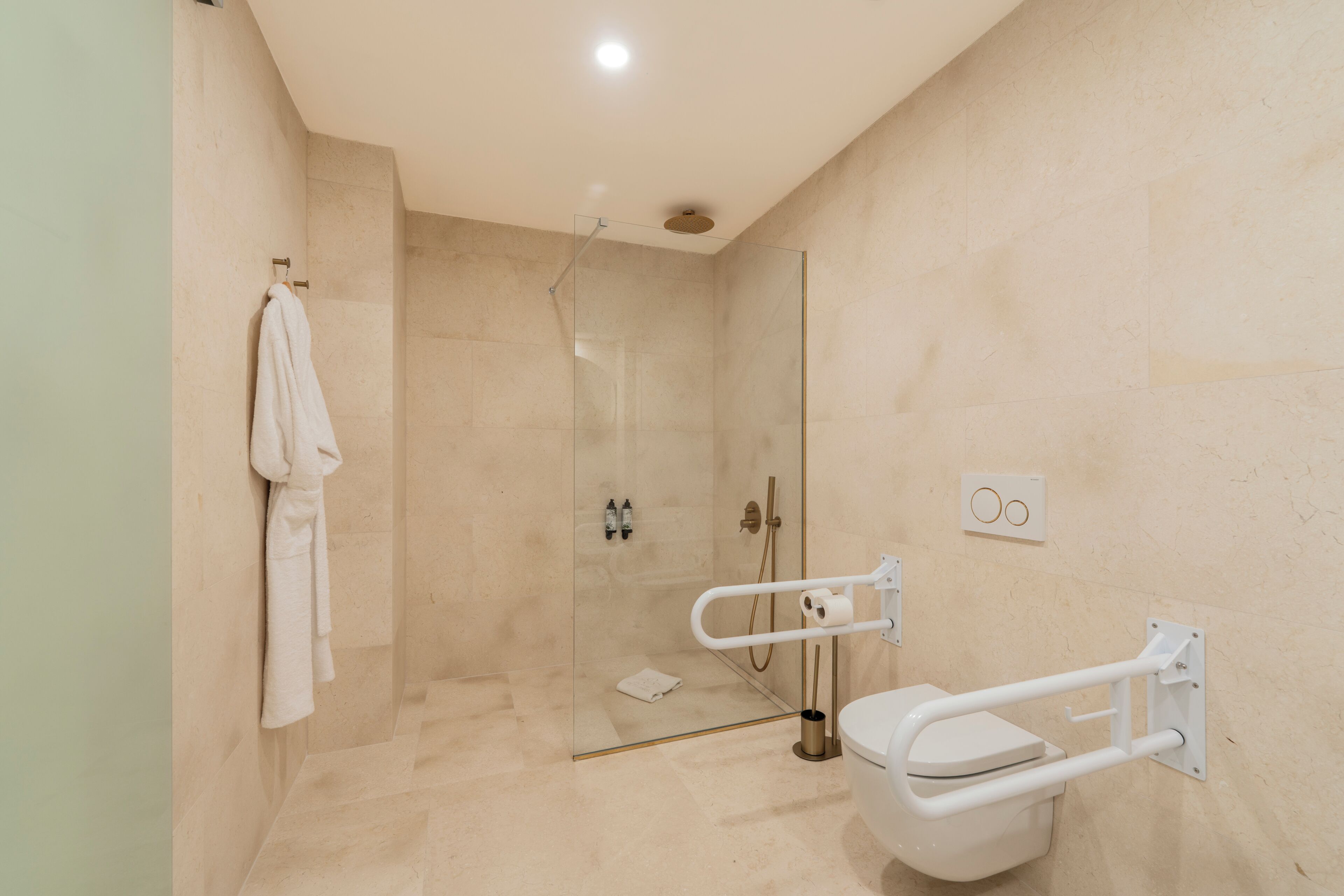 Un baño moderno con ducha envuelta en vidrio, inodoro blanco con barra de apoyo y perchero.