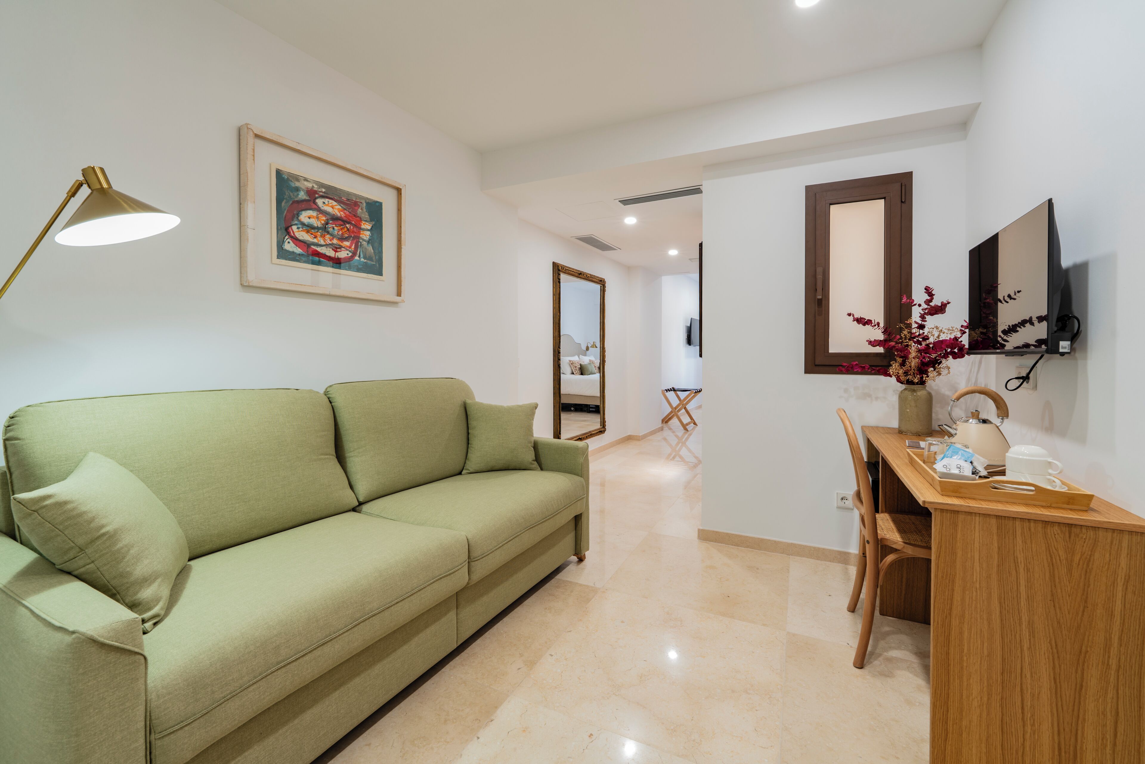 Una habitación con un sofá verde, un escritorio con silla, una lámpara, un cuadro enmarcado, un espejo y un televisor en la pared.
