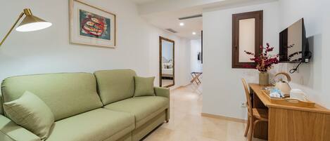 Una habitación con un sofá verde, un escritorio con silla, una lámpara, un cuadro enmarcado, un espejo y un televisor en la pared.