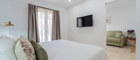 Un dormitorio con cama, televisor montado en la pared, ventana con cortinas y un sofá en la habitación contigua.