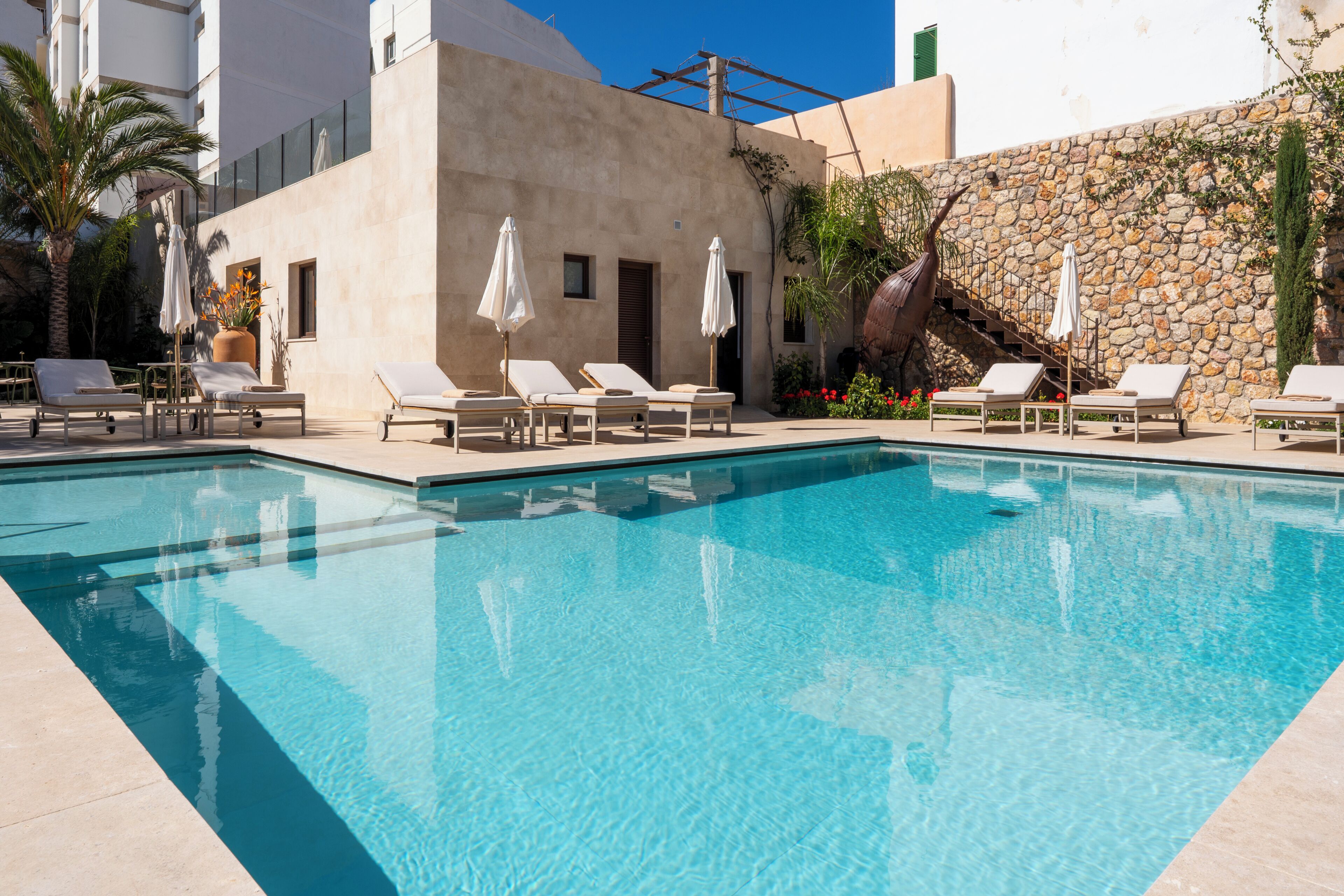 A swimming pool with sun loungers and umbrellas, surrounded by a stone wall and a building with a modern design.
