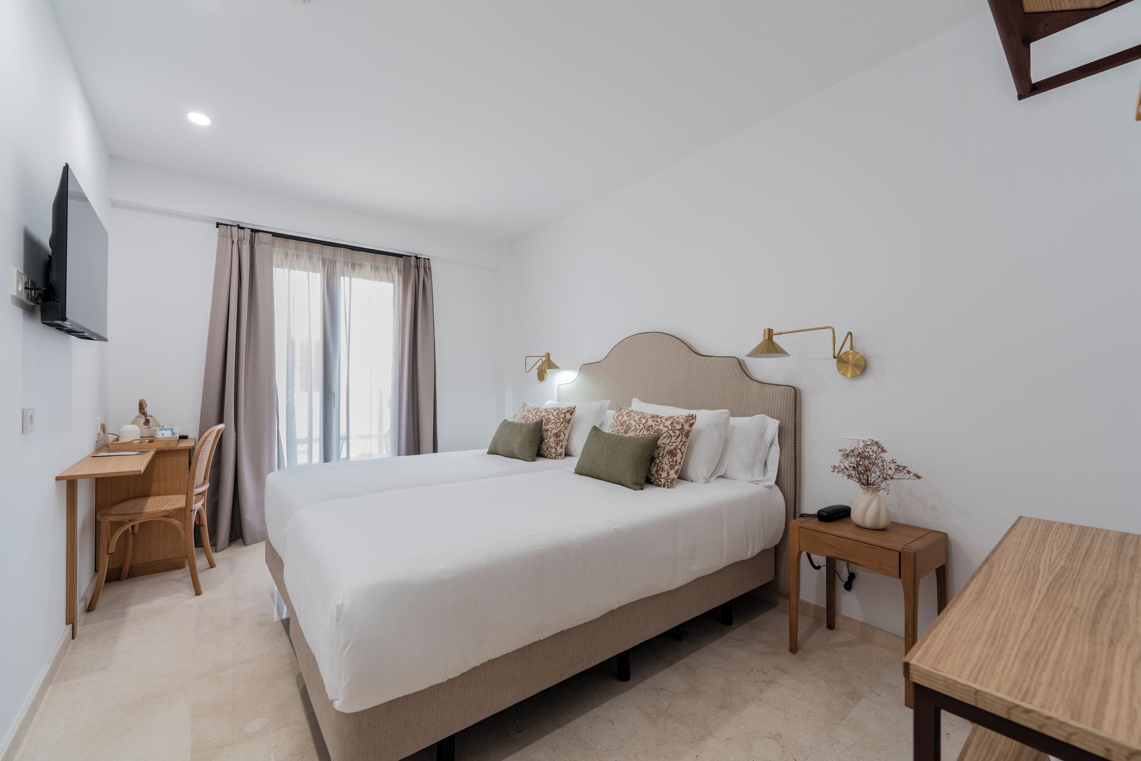 Una habitación de hotel con una cama grande, un escritorio, una silla, un televisor, una mesita de noche con lámpara y una ventana con cortinas.