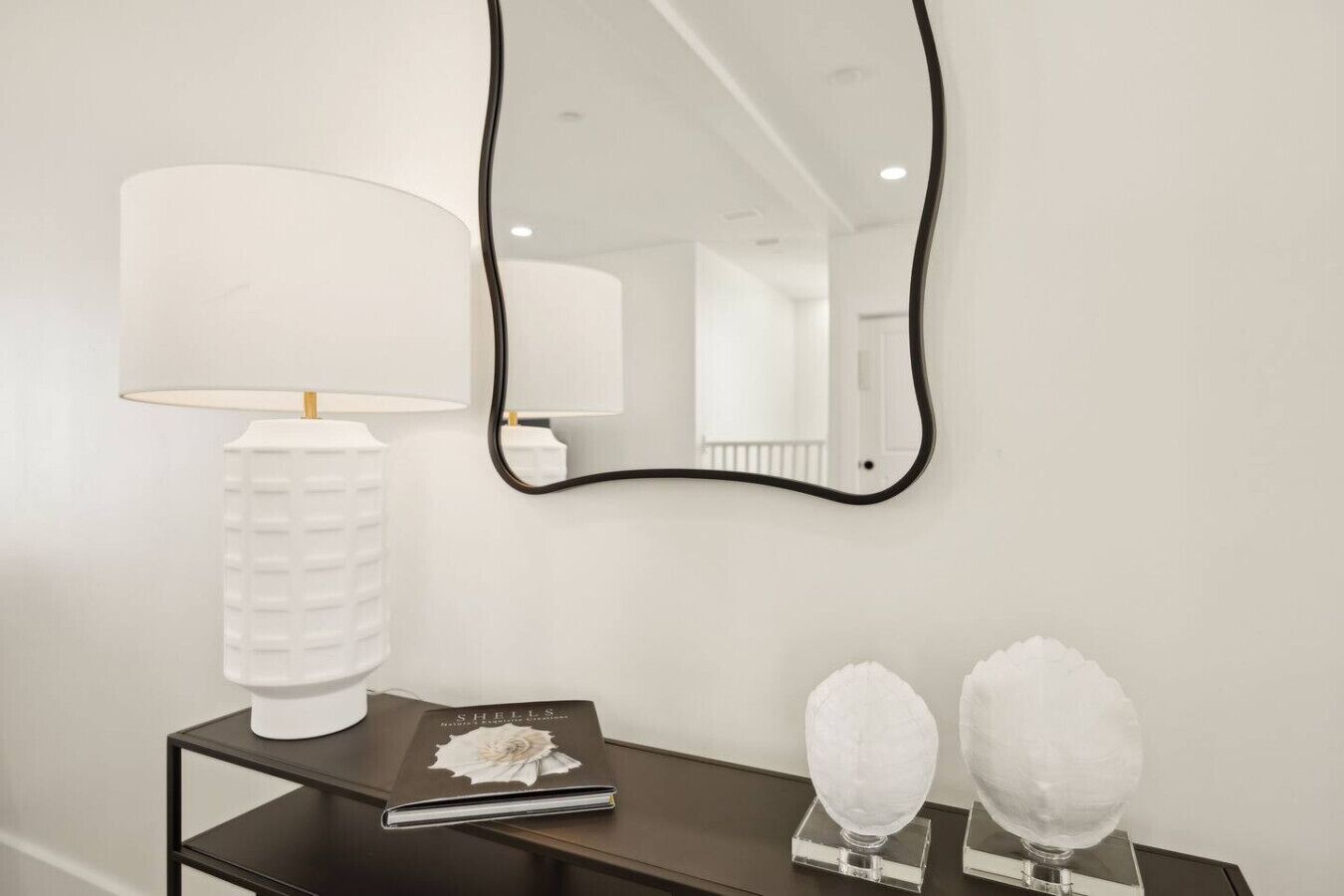 A white wall with a black-framed mirror, a white lamp, a book titled 'Shells', and two white spherical objects on a black table.