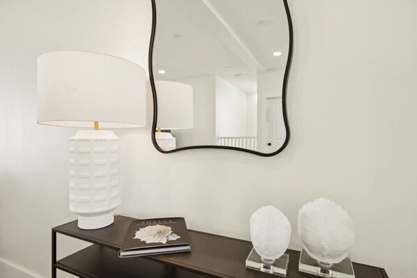 A white wall with a black-framed mirror, a white lamp, a book titled 'Shells', and two white spherical objects on a black table.