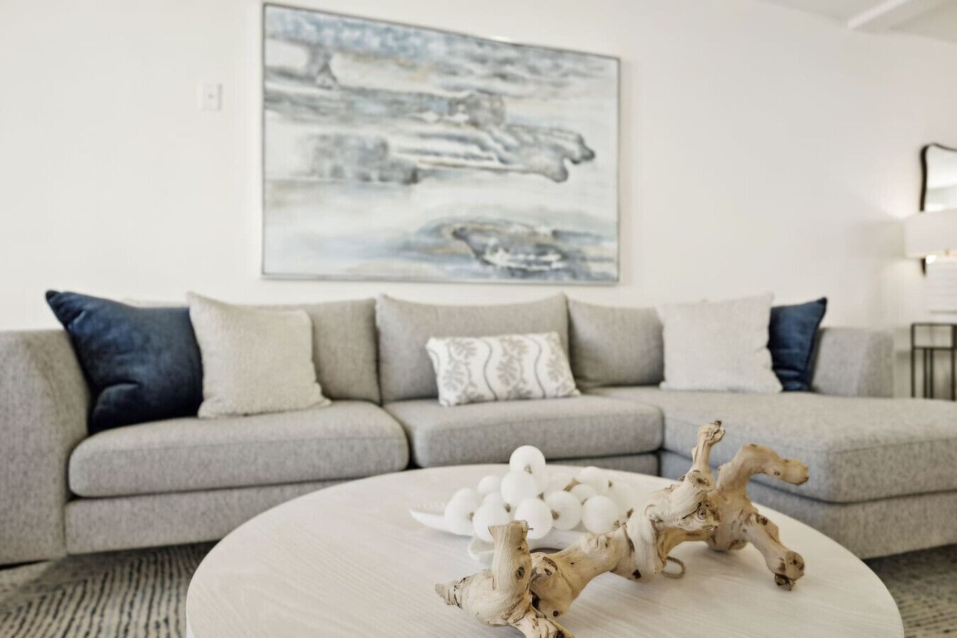 A modern living room with a grey sectional sofa, a round coffee table with decorative items, and a large abstract painting on the wall.
