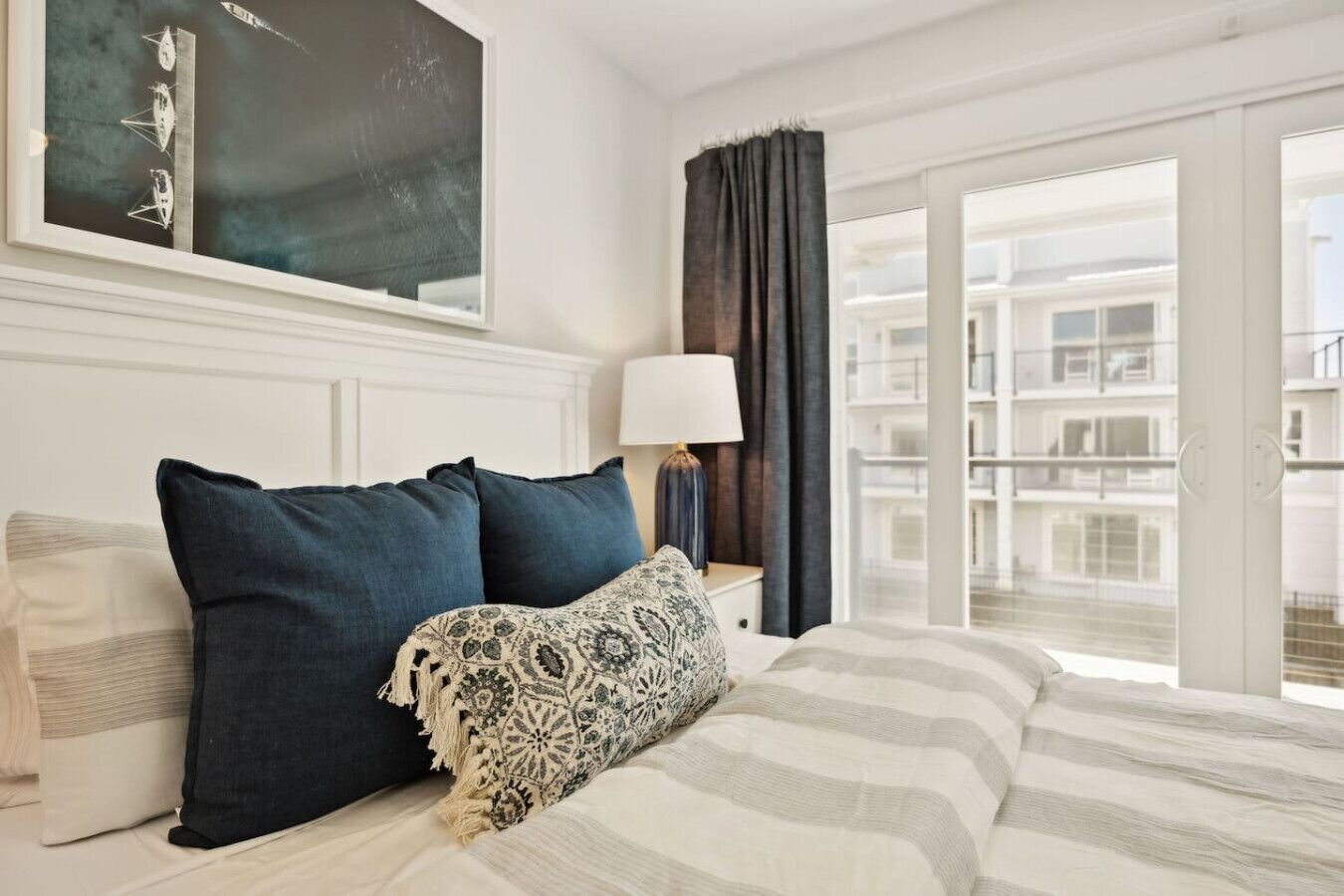 A hotel room with a bed, a nightstand, a lamp, a window with curtains, and a view of buildings.