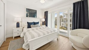 A bedroom with a large bed, white nightstands, a ceiling fan, and a view of buildings through a sliding glass door.