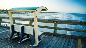 A wooden pier with a bench and a covered seating area overlooking a beach with people and waves.