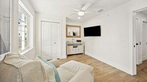 A spacious living room with a large window, a ceiling fan, a flat-screen TV, and a white cabinet.