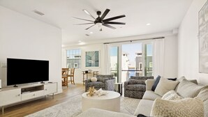 Un salon moderne doté d’un téléviseur à écran plat, d’un ventilateur de plafond et d’une grande fenêtre offrant une vue sur les bâtiments.
