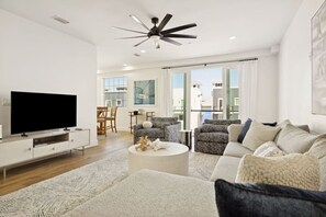 A modern living room with a flat-screen TV, a ceiling fan, and a large window offering a view of buildings.