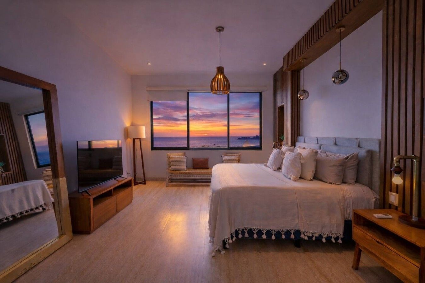 A bedroom with a large bed, a television, a window with a sunset view, and a mirror.