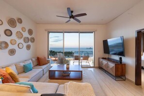 A spacious living room with a large TV, a ceiling fan, and a view of the sea through large windows. - Above The Arch: Penthouse w/ Private Rooftop Pool (Cabo San Lucas)