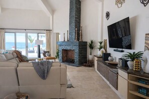 A living room with a stone fireplace, a sofa, a TV, and a view of the ocean. - The Beachhouse At Rancho Leonero (Los Cabos)