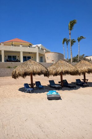 Ein Strand mit reetgedeckten Sonnenschirmen und Liegestühlen, ein Gebäude mit rotem Dach und Palmen im Hintergrund.