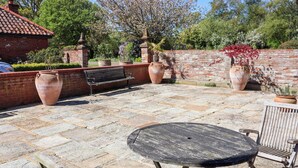 A patio with stone flooring, a round table, wooden chairs, a bench, and large planters. - Winston Lodge (Beccles)
