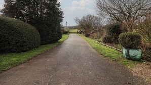 A rural road flanked by well-maintained hedges and trees, leading into a distant field. - The Gun Room (Beccles)