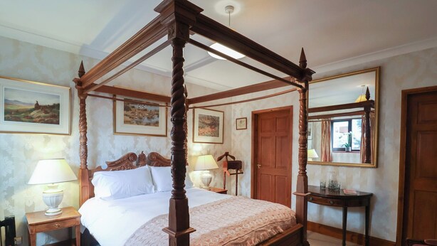 A four-poster bed with a wooden frame, a bedside table with a lamp, a nightstand, and a chair. - The Gun Room (Beccles)