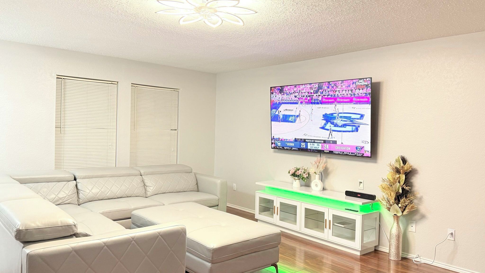 A modern living room with a sectional sofa, a flat-screen TV mounted on the wall, a white cabinet with glass doors, and a small table with decorative items.