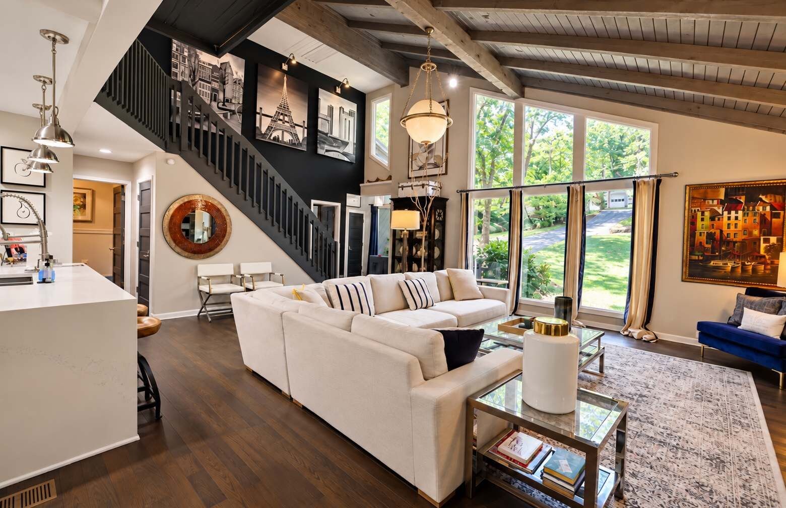 A spacious living area with a large white sofa, a glass coffee table, and a blue sofa. A staircase with a black railing leads to an upper level.