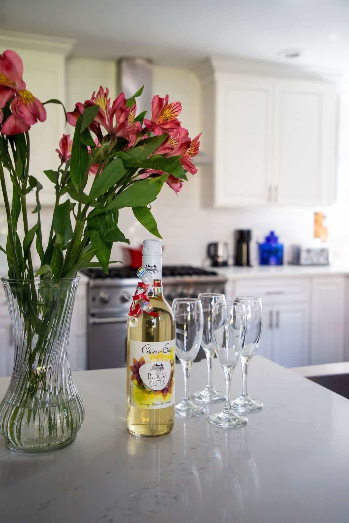 A bottle of wine, a vase with flowers, and empty wine glasses on a kitchen counter.