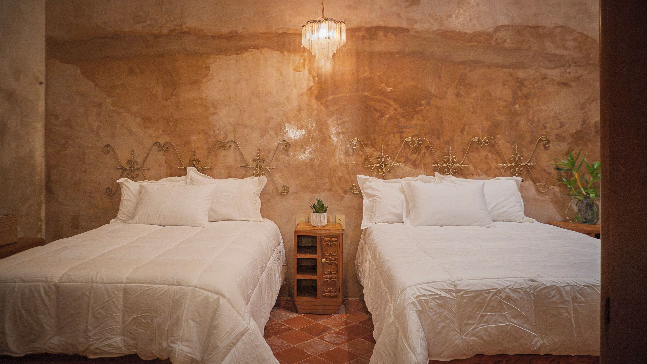 A room with two beds, a small wooden nightstand with a plant, and a wall-mounted light fixture.