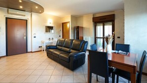 A living room with a leather sofa, a wooden dining table with chairs, a small white side table, and a large window with a view. - Milano Certosa Apartment (Milano)