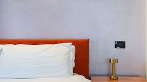 A bed with white bedding and an orange headboard, a bedside table with a gold lamp, and a wall-mounted control panel. - Milano Certosa Apartment (Milano)