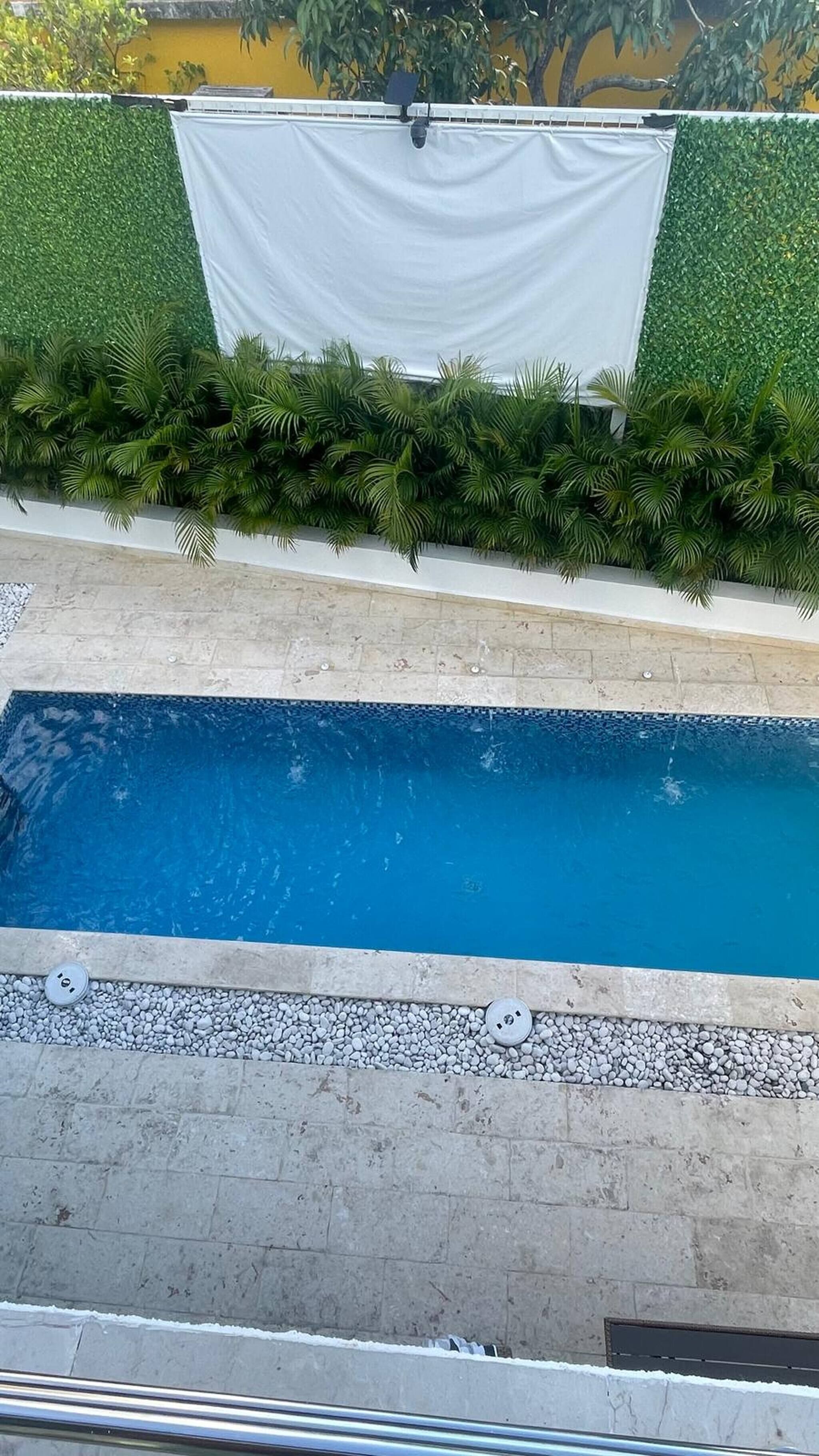 Ein Schwimmbecken mit weißem Rand, umgeben von einer niedrigen Hecke und einem weißen Stoff, der über einer Struktur drapiert ist.