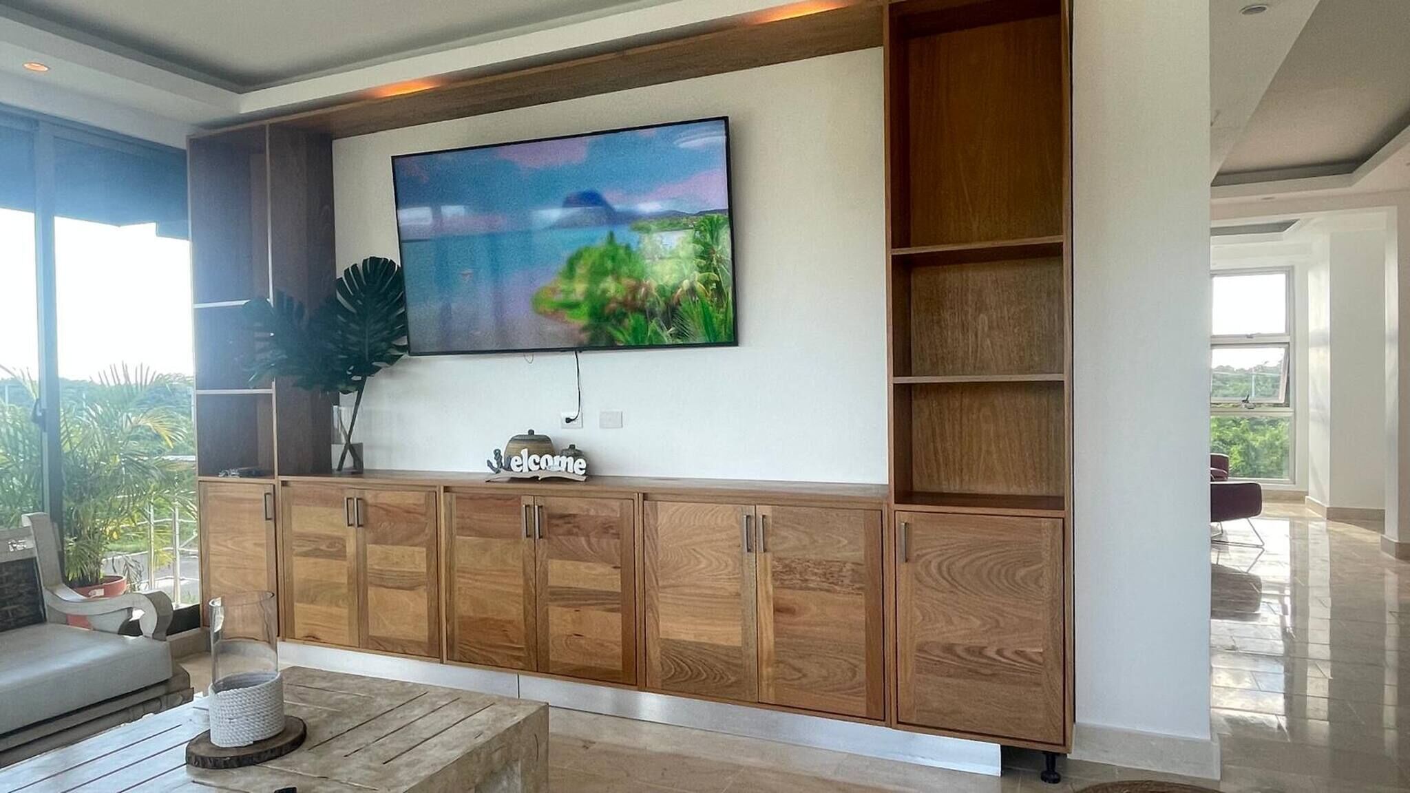 Una sala de estar moderna con un televisor de pantalla plana grande montado sobre un mueble de madera, un espacio de descanso cómodo con un sofá y una mesa de centro, y un amplio ventanal con vistas a la vegetación exterior.