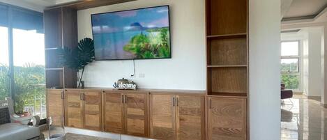 Una sala de estar moderna con un televisor de pantalla plana grande montado sobre un mueble de madera, un espacio de descanso cómodo con un sofá y una mesa de centro, y un amplio ventanal con vistas a la vegetación exterior.