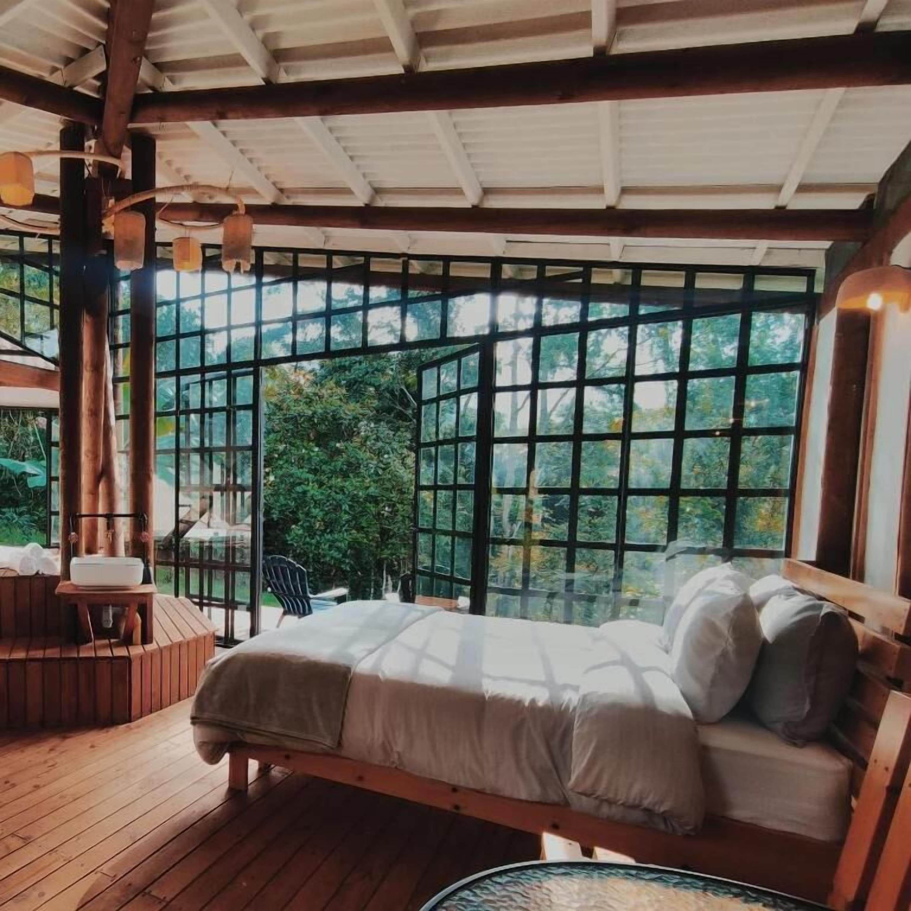 Una cama de madera con vistas a la vegetación a través de ventanas amplias, una mesita pequeña y una silla.