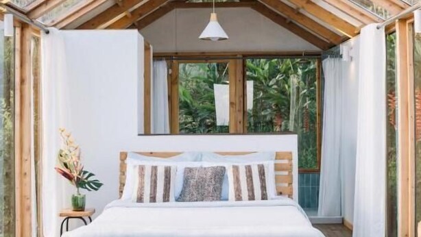 Un dormitorio acogedor con cama de madera, lámpara colgante y ventanas amplias que dan a vistas de vegetación exterior.